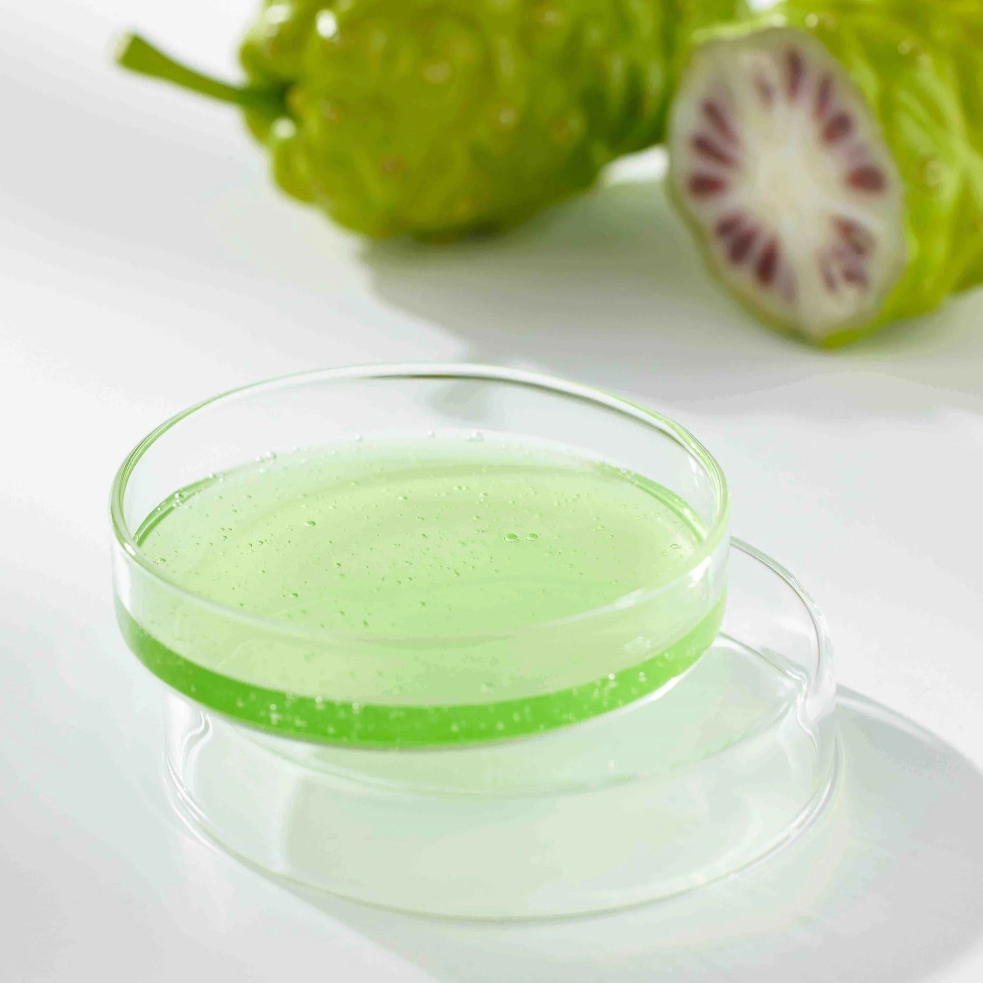 CELIMAX Noni Ampoule Calming+Radiance 100ml in a clear dish with sliced noni fruit in the background