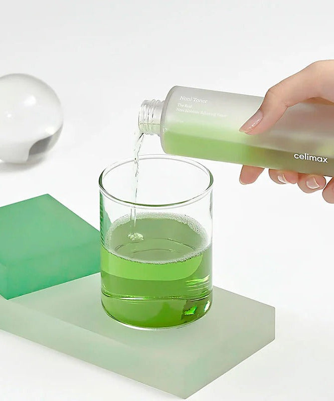 CELIMAX The Real Noni Moisture Balancing Toner 320ml being poured into a glass with green fluid, showcasing product texture.