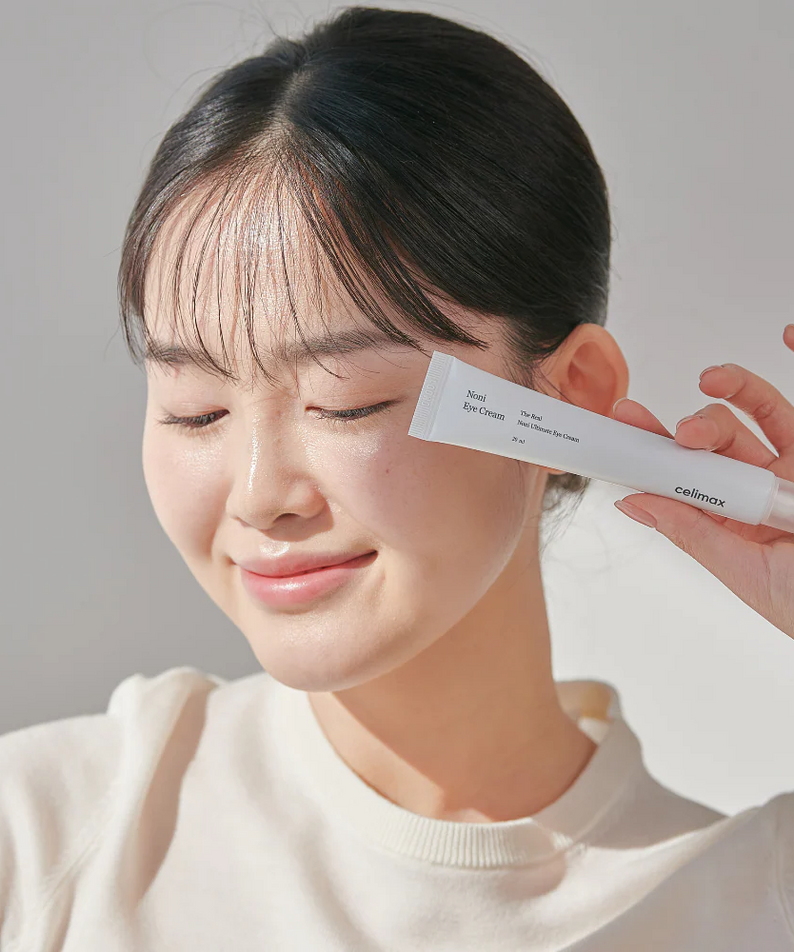 Woman holding CELIMAX The Real Noni Ultimate Eye Cream 20ml with a smile, showcasing skincare product.