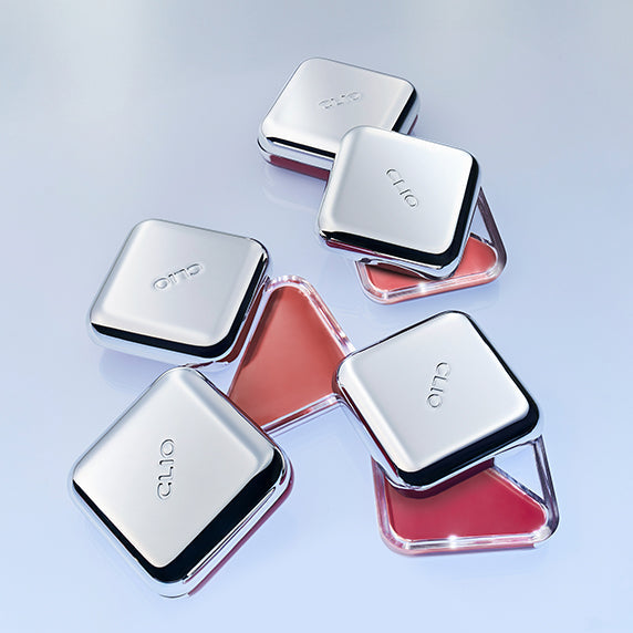 Open and closed silver compact cases of CLIO Essential Lip Cheek showcasing a blend for lip and cheek makeup on a reflective surface.