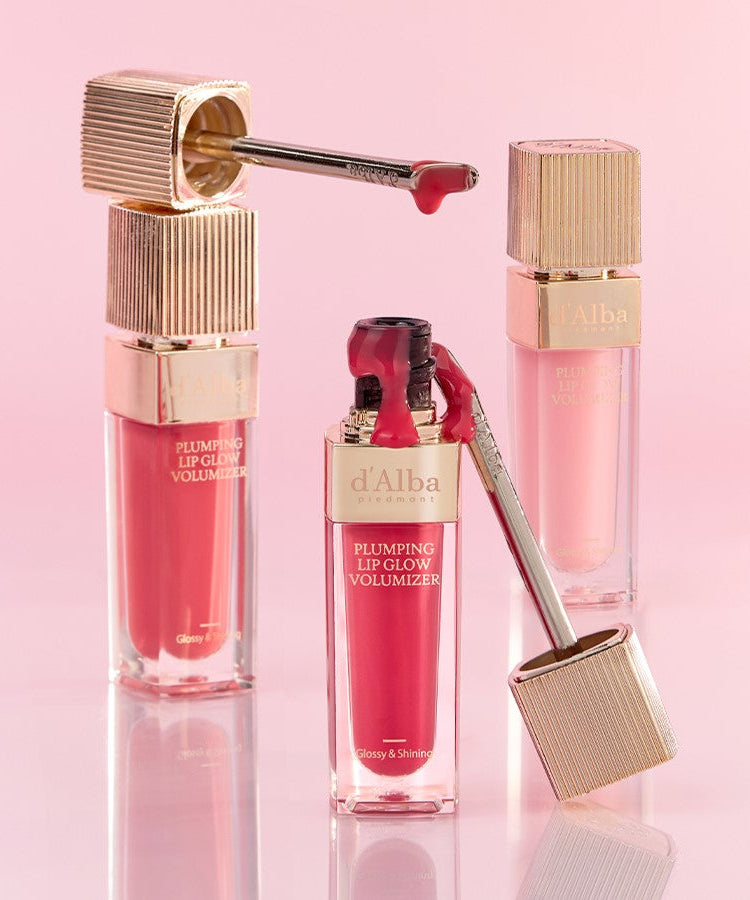 D'ALBA Plumbing Lip Glow Mood Volumizer in pink and red shades with gold accents, set against a soft pink background.