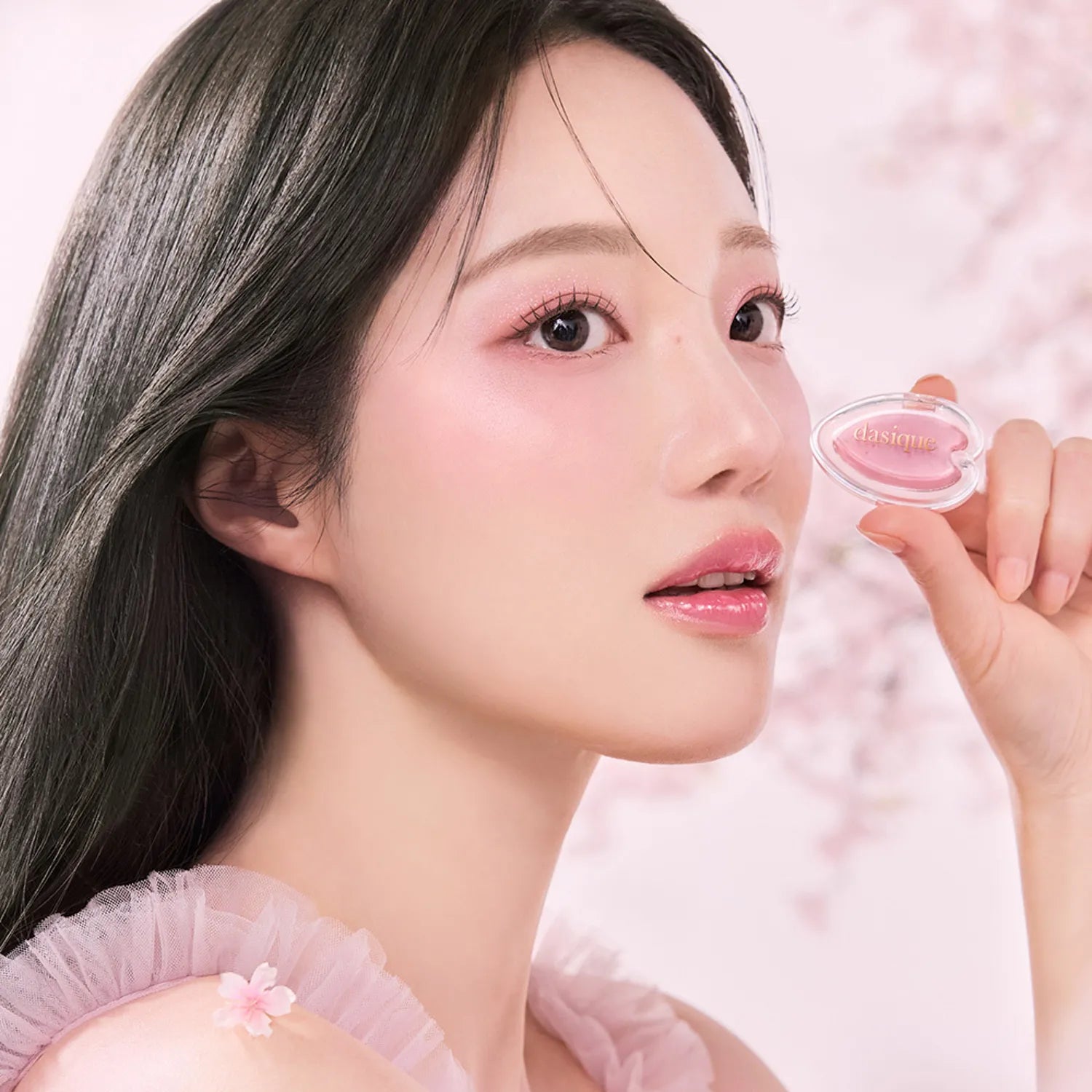 Woman holding DASIQUE Blooming Single Shadow with pink floral background, highlighting soft makeup look.