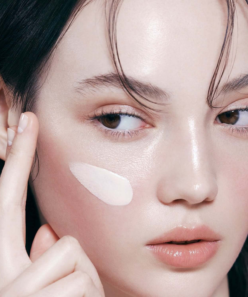 Model applying DEAR DAHLIA Prime Layer Glow Primer on cheek, showcasing its smooth texture and luminous finish for radiant skin.