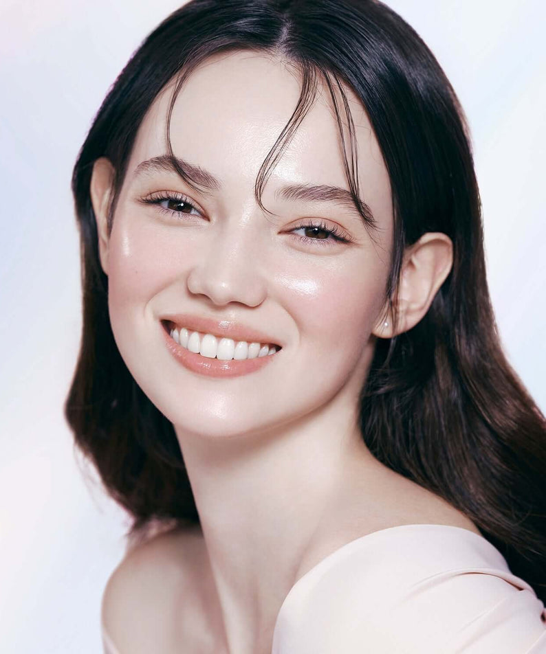 Woman with radiant skin wearing DEAR DAHLIA Prime Layer Glow Primer, showcasing makeup benefits.