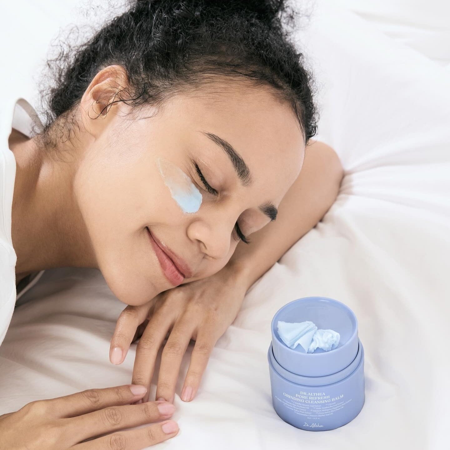 Woman applying DR.ALTHEA Pore Refresh Grinding Cleansing Balm 50ml on face while relaxing on bed