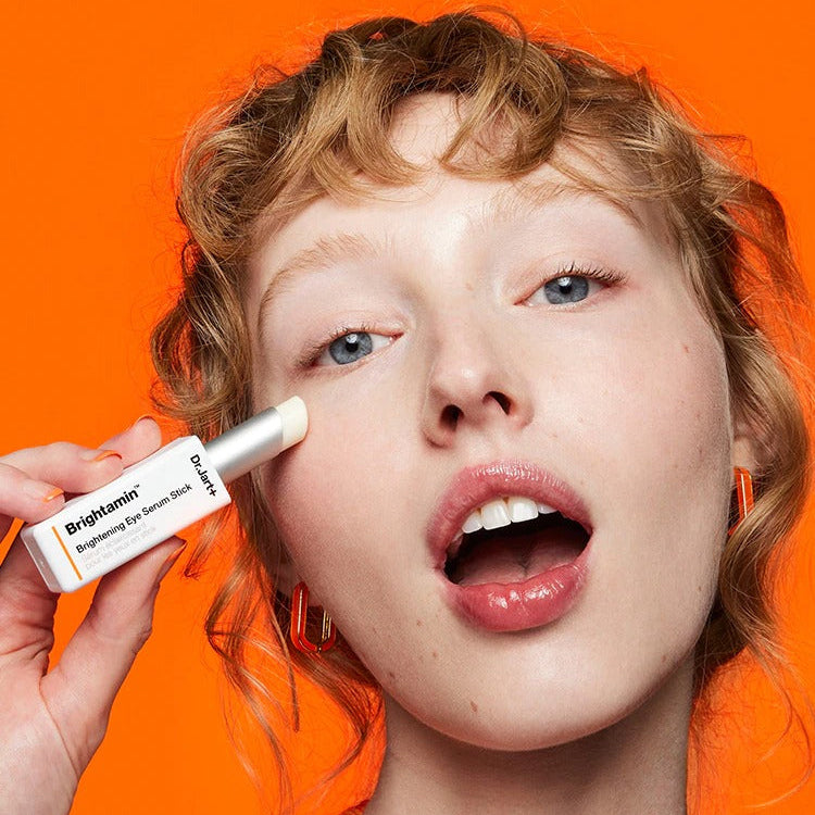 Person using DR.JART+ Brightamine Brightening Eye Serum Stick against vibrant orange background.