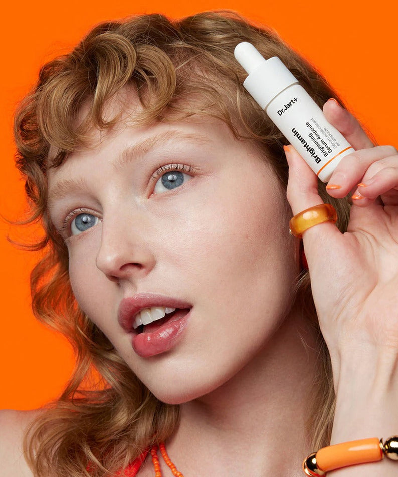 Woman holding DR.JART+ Brightamine Brightening Serum Ampoule against orange background