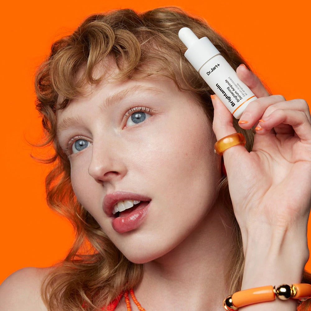 Woman holding DR.JART+ Brightamine Brightening Serum Ampoule against orange background
