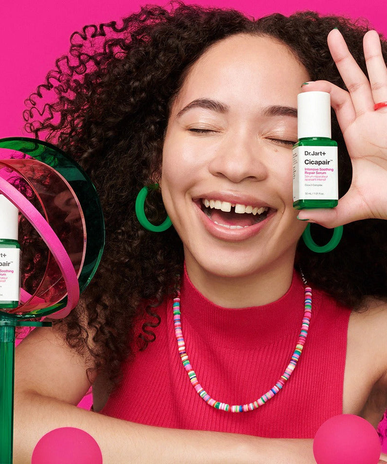 Smiling woman holding DR.JART+ CicapairI ntensive Soothing Repair Serum 30ml against pink background.