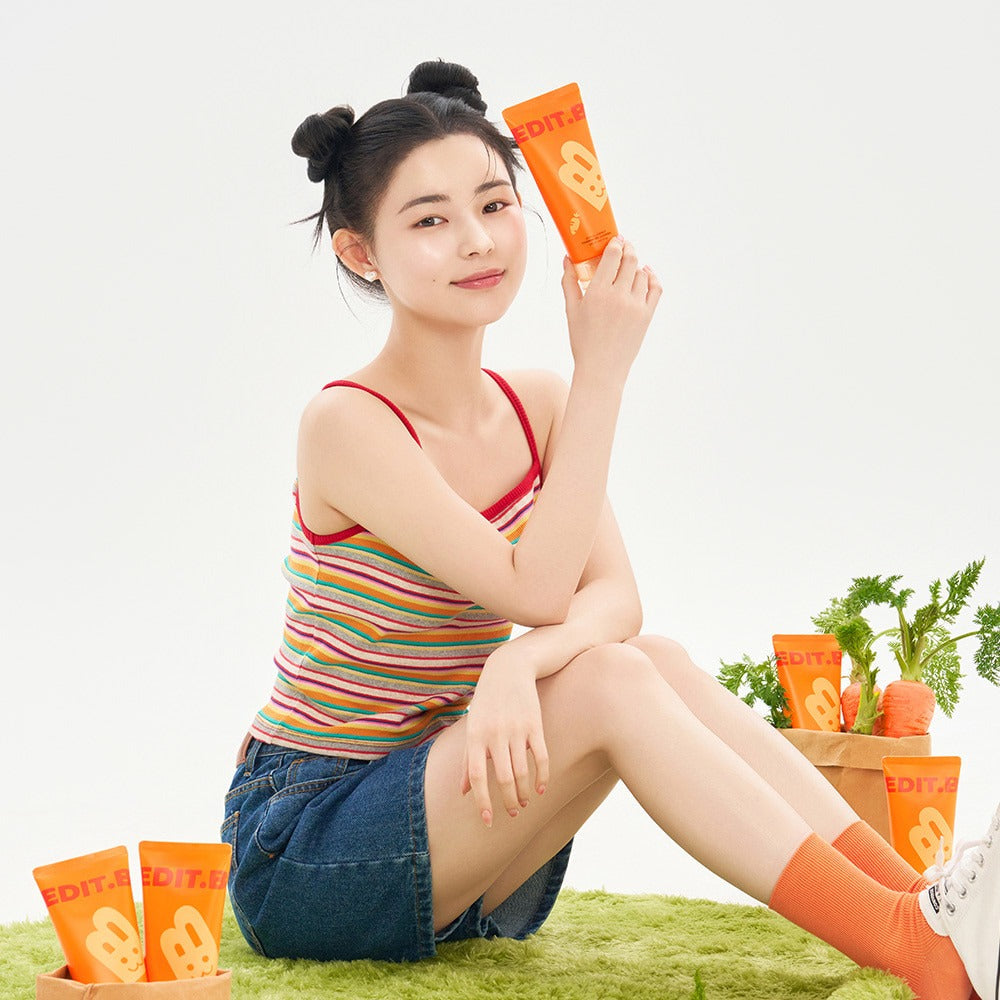 Person holding EDIT.B Joyful Carrot Foaming Gel Cleanser 150ml with orange packaging, surrounded by product tubes and fresh carrots.