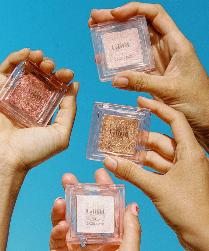 Hands holding GLINT Glint Highlighter (Marble) against a blue sky background, showcasing the product's four shimmering shades.