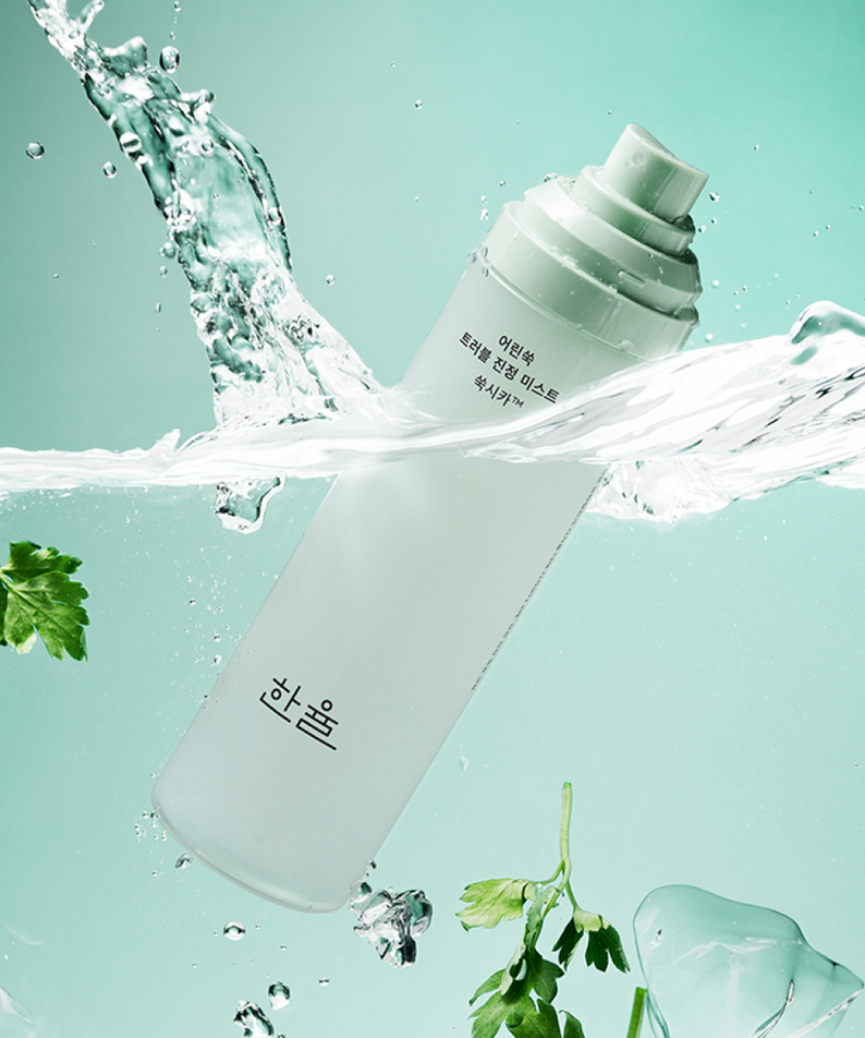 HANYUL Young Artemisia Trouble Mist 120ml bottle surrounded by fresh green herbs and splashing water on a teal background.