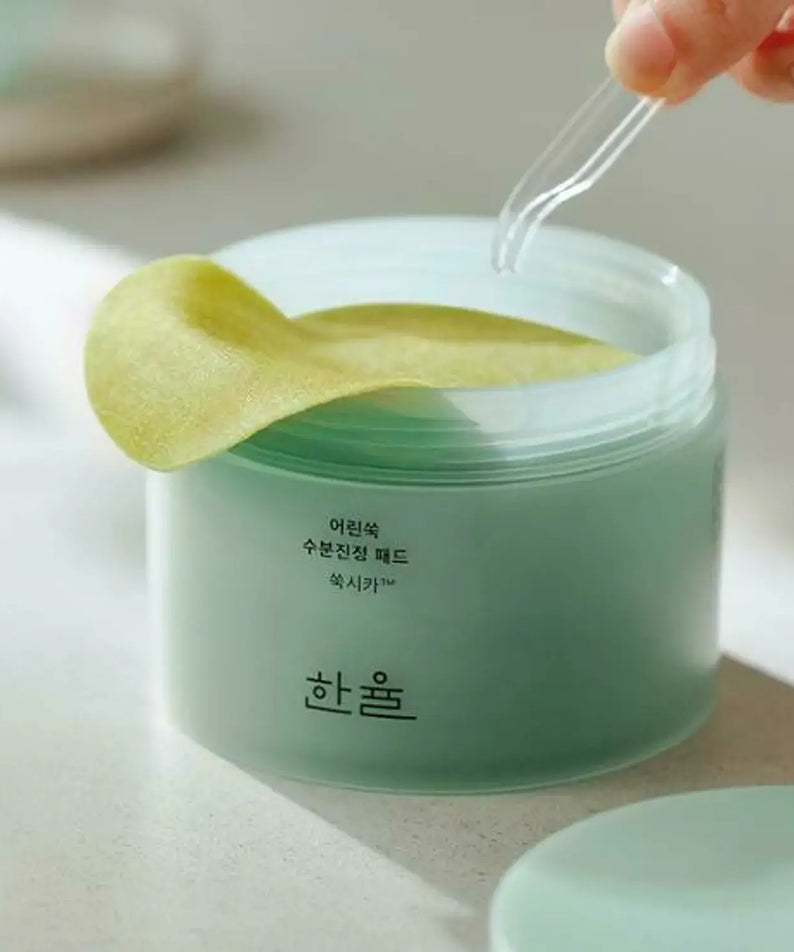 Close-up of HANYUL Young Mugwort Moisturizing Soohting Pad container with pads being picked up by tweezers, showing product texture.