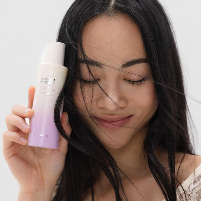 Woman holding HARUHARU Black Rice Probiotics Barrier Essence 120ml in her hand, smiling with closed eyes, against a light background.