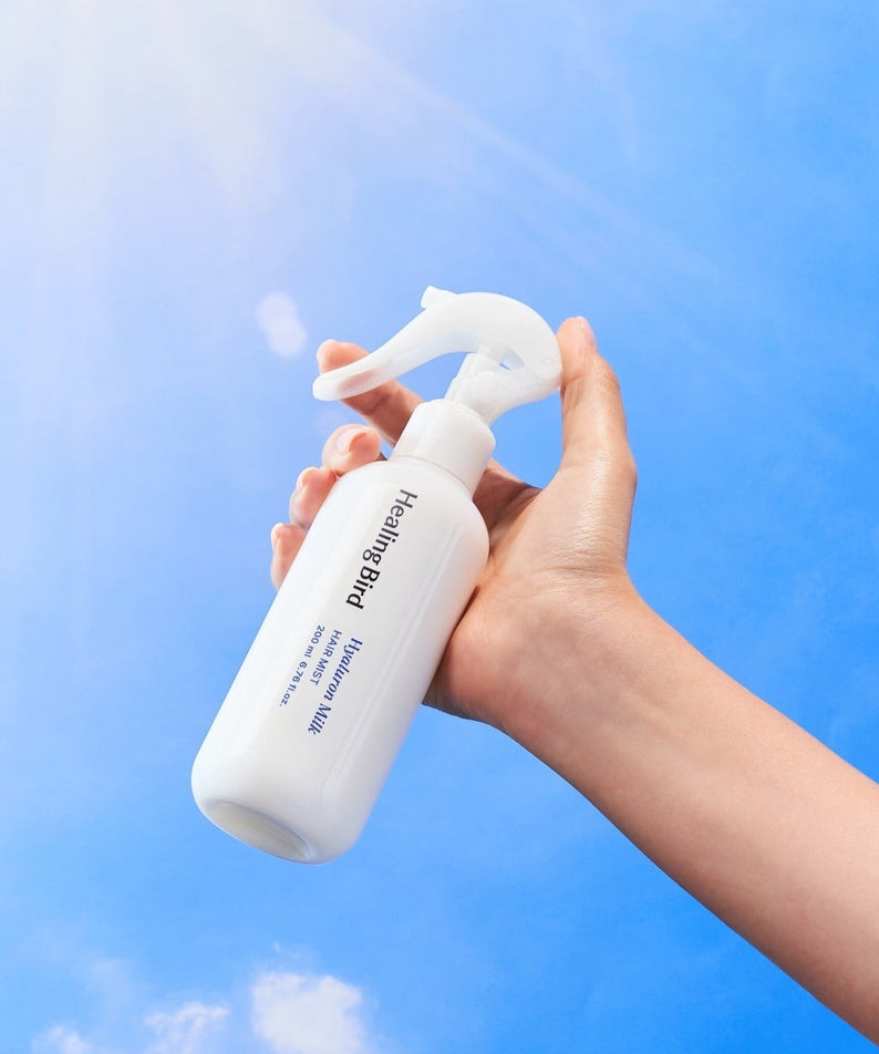 Hand holding HEALING BIRD Hyalutonic Milk Hair Mist 200ml against blue sky background.