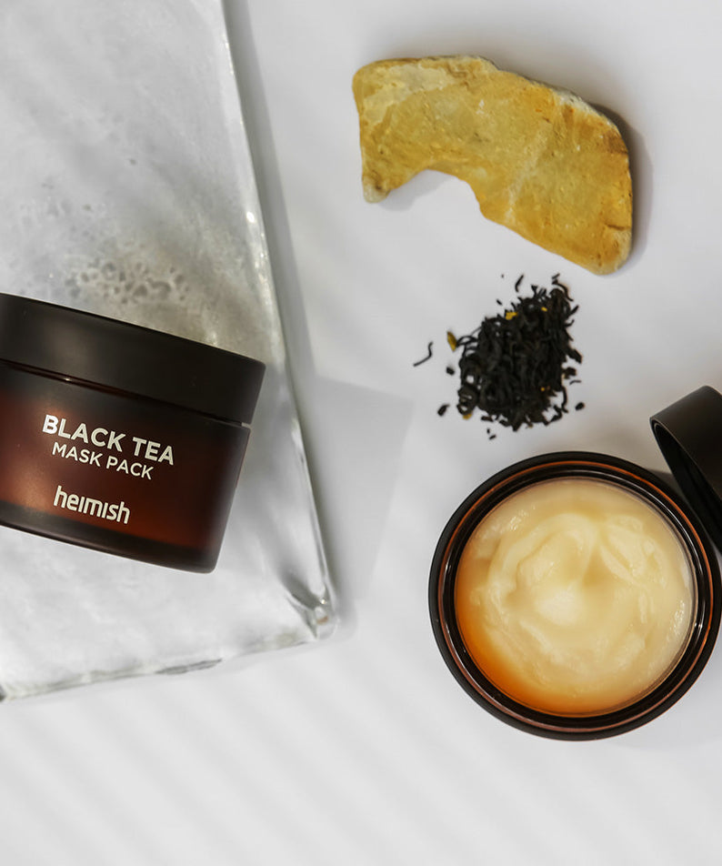 HEIMISH Black Tea Mask Pack 110ml jar with contents on display next to tea leaves and rock, showcasing skincare product.