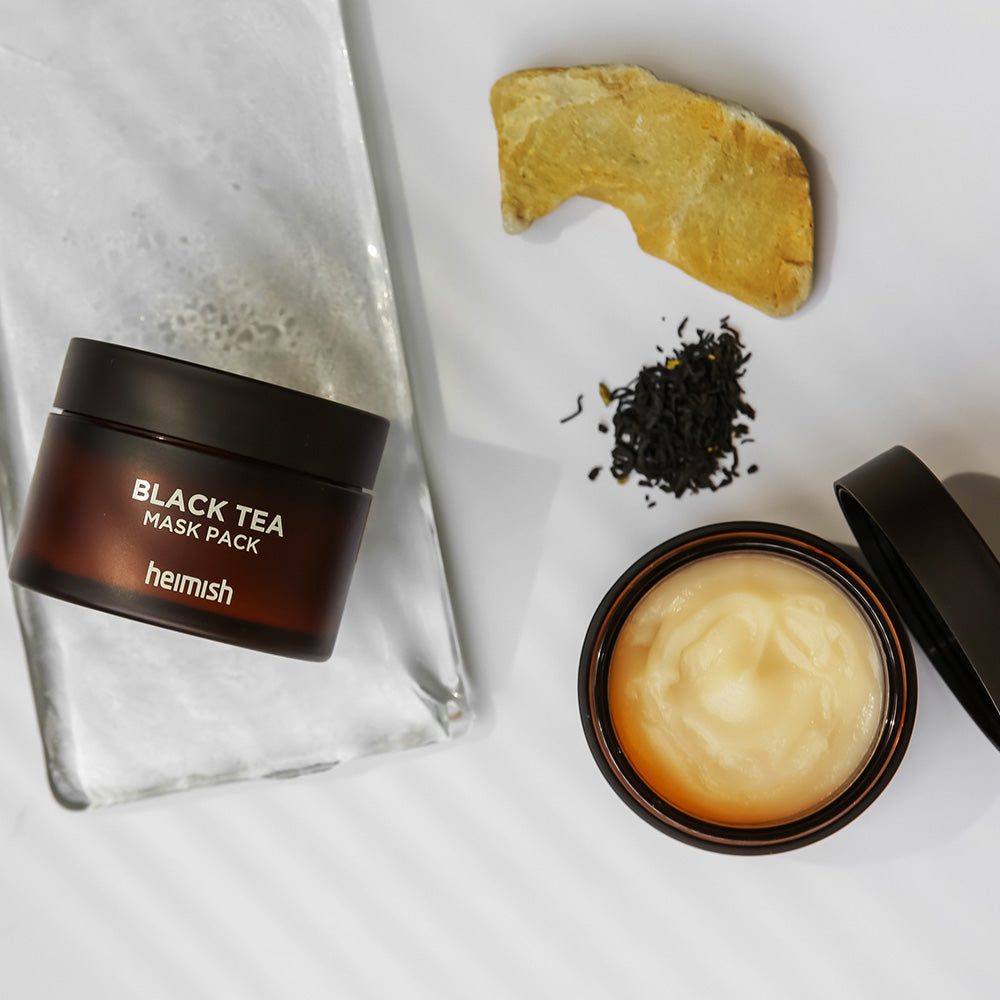 HEIMISH Black Tea Mask Pack 110ml jar with contents on display next to tea leaves and rock, showcasing skincare product.