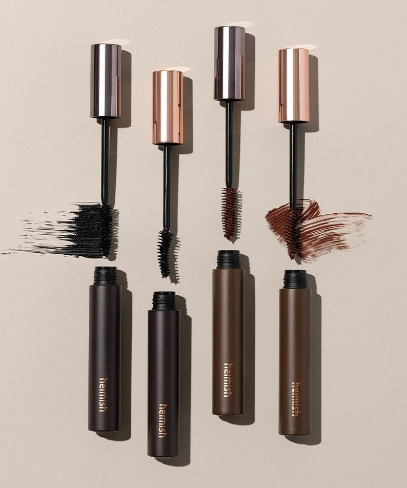 HEIMISH Dailism Mascara in black, brown, and dark brown shades, showing open tubes and brush strokes on a beige background.