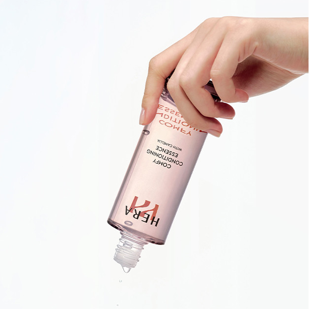 Hand holding HERA Comfy Conditioning Essence 150g bottle dripping, highlighting the product's hydrating properties.