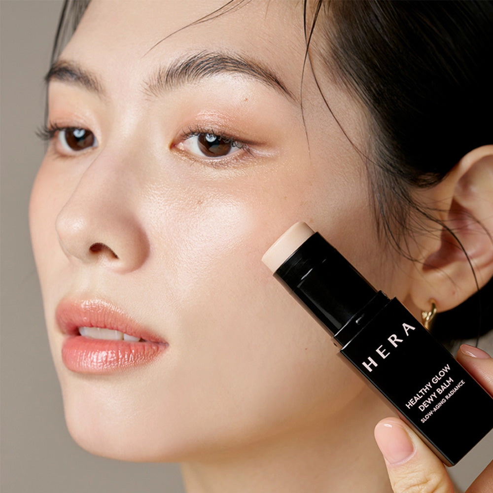 Woman applying HERA Healthy Glow Dewy Balm 10g to her cheek for a radiant look.