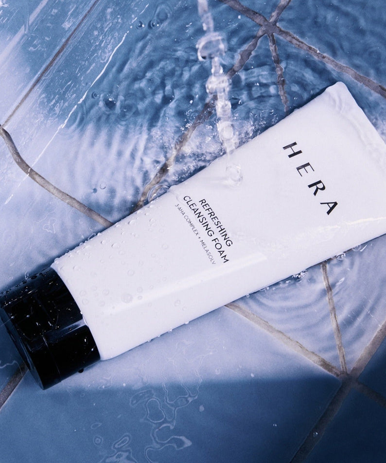 HERA Refreshing Cleansing Foam 160g tube under running water on a tiled surface, showcasing its refreshing cleansing properties.