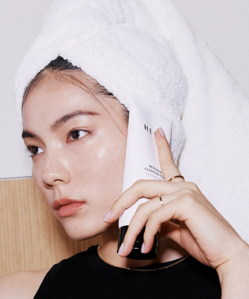 Person holding HERA Refreshing Cleansing Foam 160g next to face, showcasing skincare product.