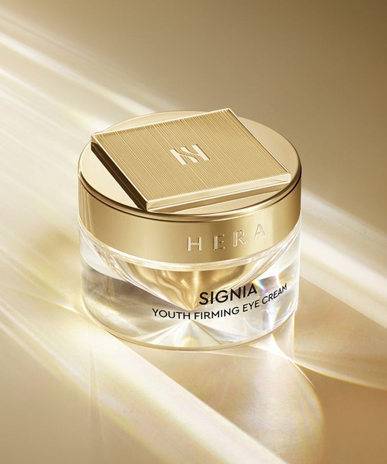HERA Signa Youth Firming Eye Cream 30ml in elegant gold packaging on a light-reflective surface.