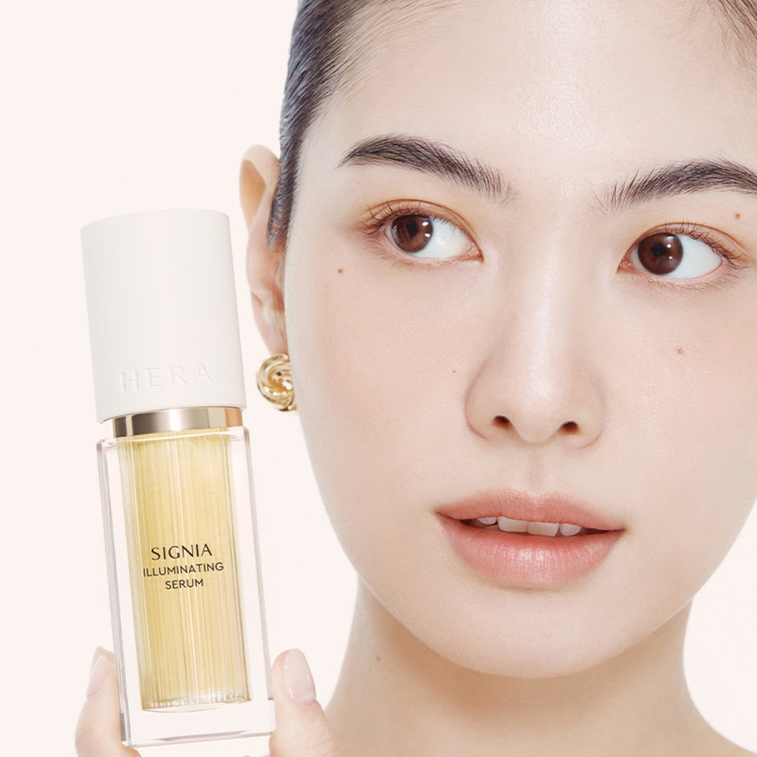 Woman holding HERA Signia Illuminating Serum 30ml with close-up facial expression.