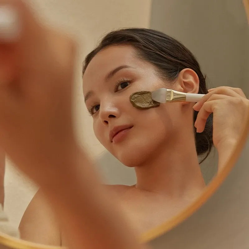 Woman applying HOLITUAL Clean Therapy Powder Mask with brush in mirror reflection
