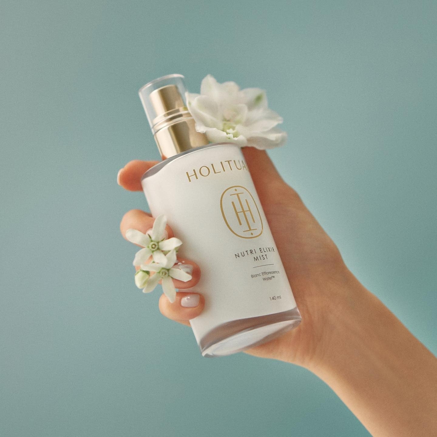 Hand holding HOLITUAL Elixir Mist 140ml bottle with flowers accentuating the design.