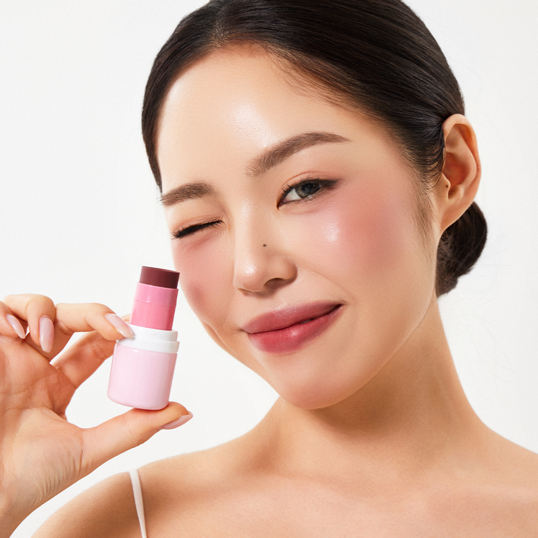 Woman holding HOUSE OF HUR Every Cheek Blush in a pink shade, smiling with soft, glowing skin.