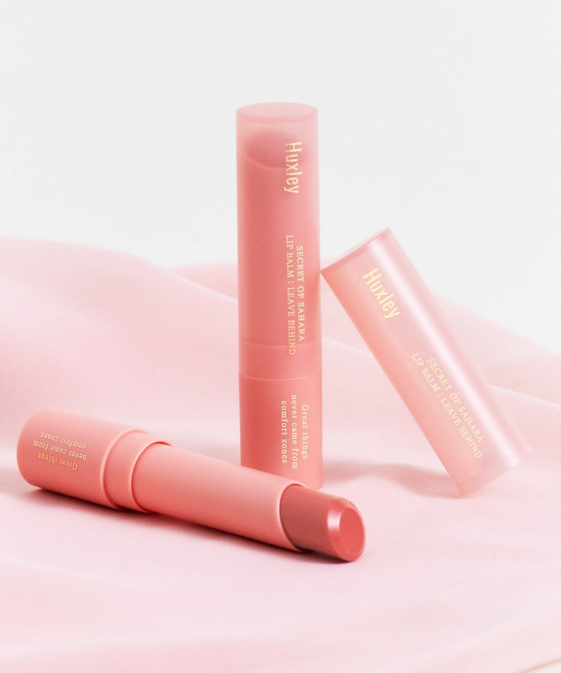 HUXLEY Lip Balm Leave Behide on pink fabric backdrop, showcasing three tubes of the product.
