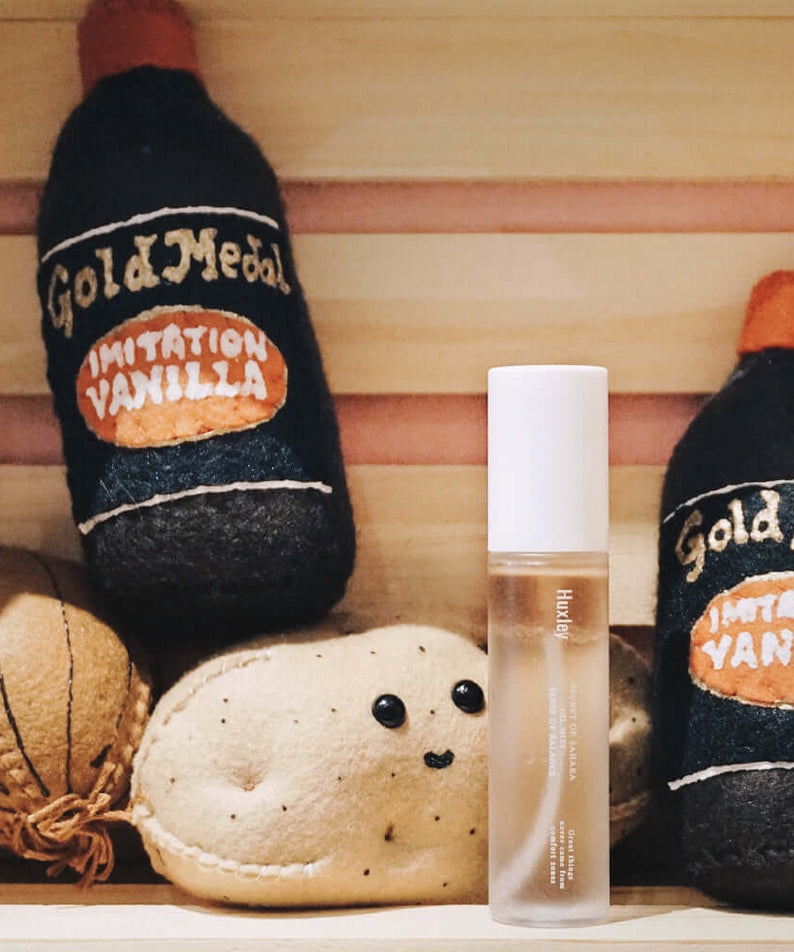 HUXLEY Oil Mist Sense Of Balance displayed with vintage vanilla bottles and plush toy in a wooden shelf scene.