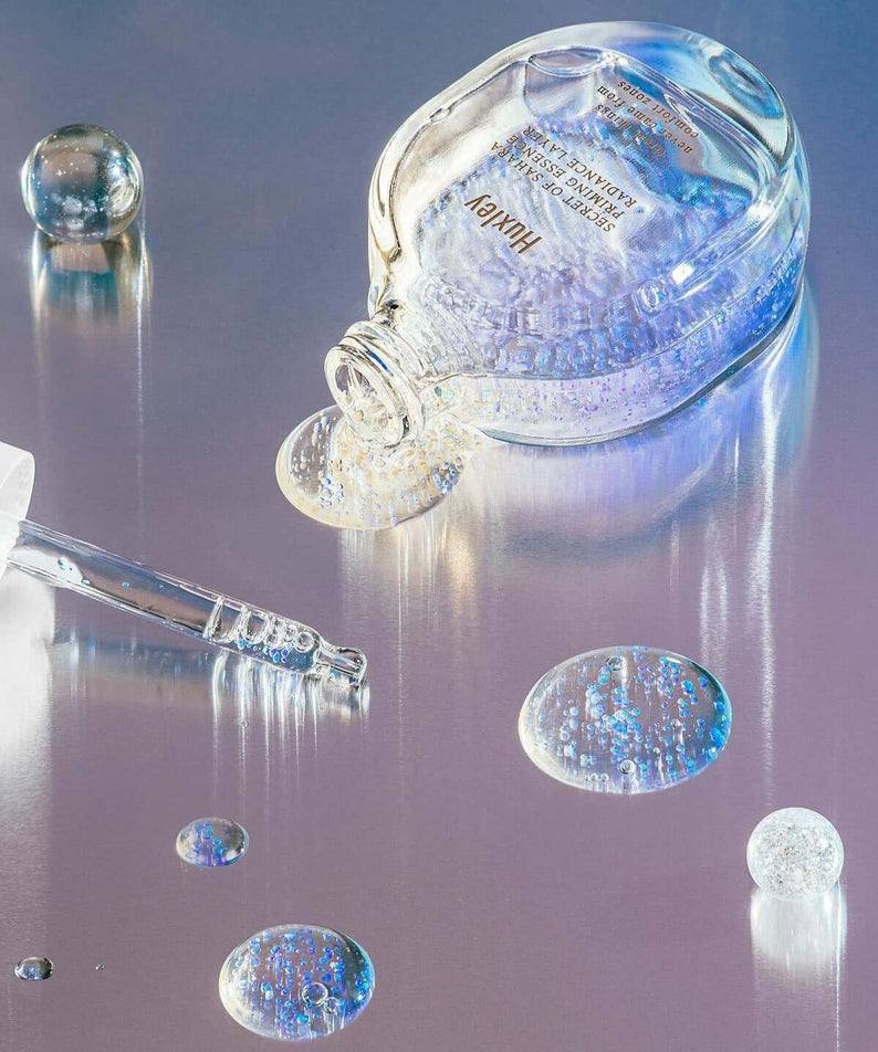 HUXLEY Priming Essence Rediance Layer bottle and dropper with iridescent liquid on metallic surface, highlighting radiant skincare.