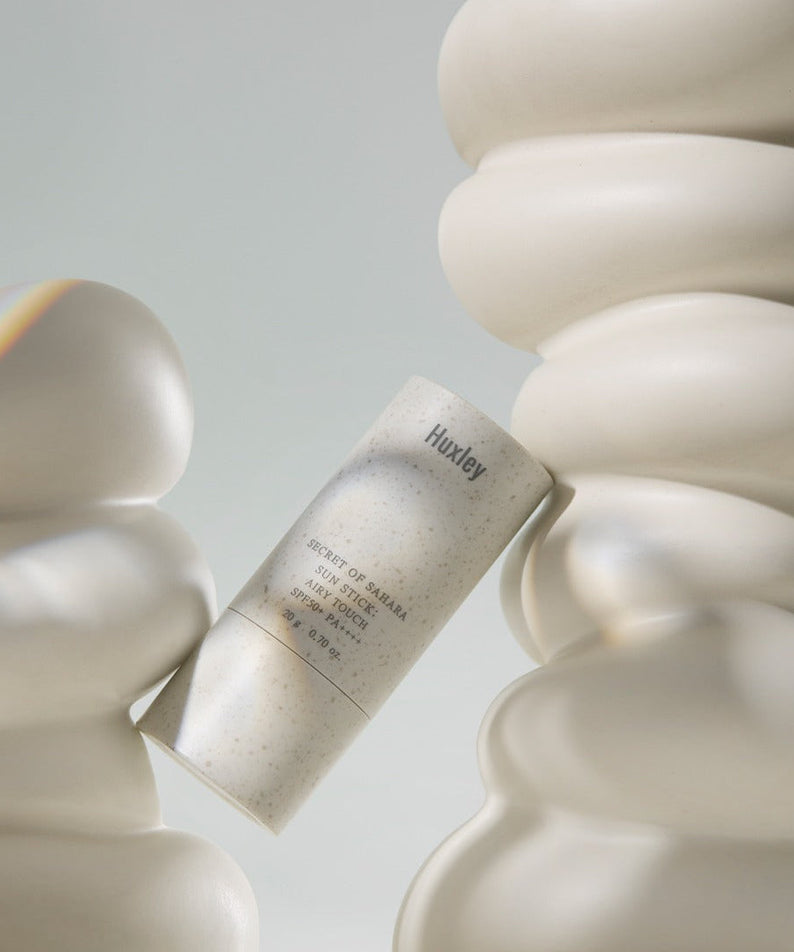 HUXLEY Sun Stick Airy Touch 20g in elegant packaging, surrounded by abstract shapes, offering UV protection and skincare benefits.