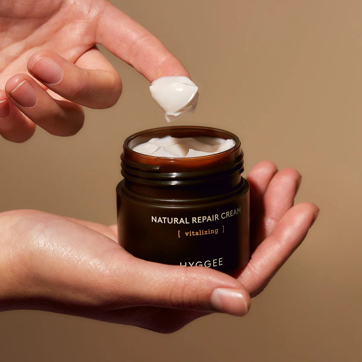 Close-up of hands holding HYGGEE Nature Repair Cream 50ml jar with cream on fingertip, showcasing its texture and packaging.