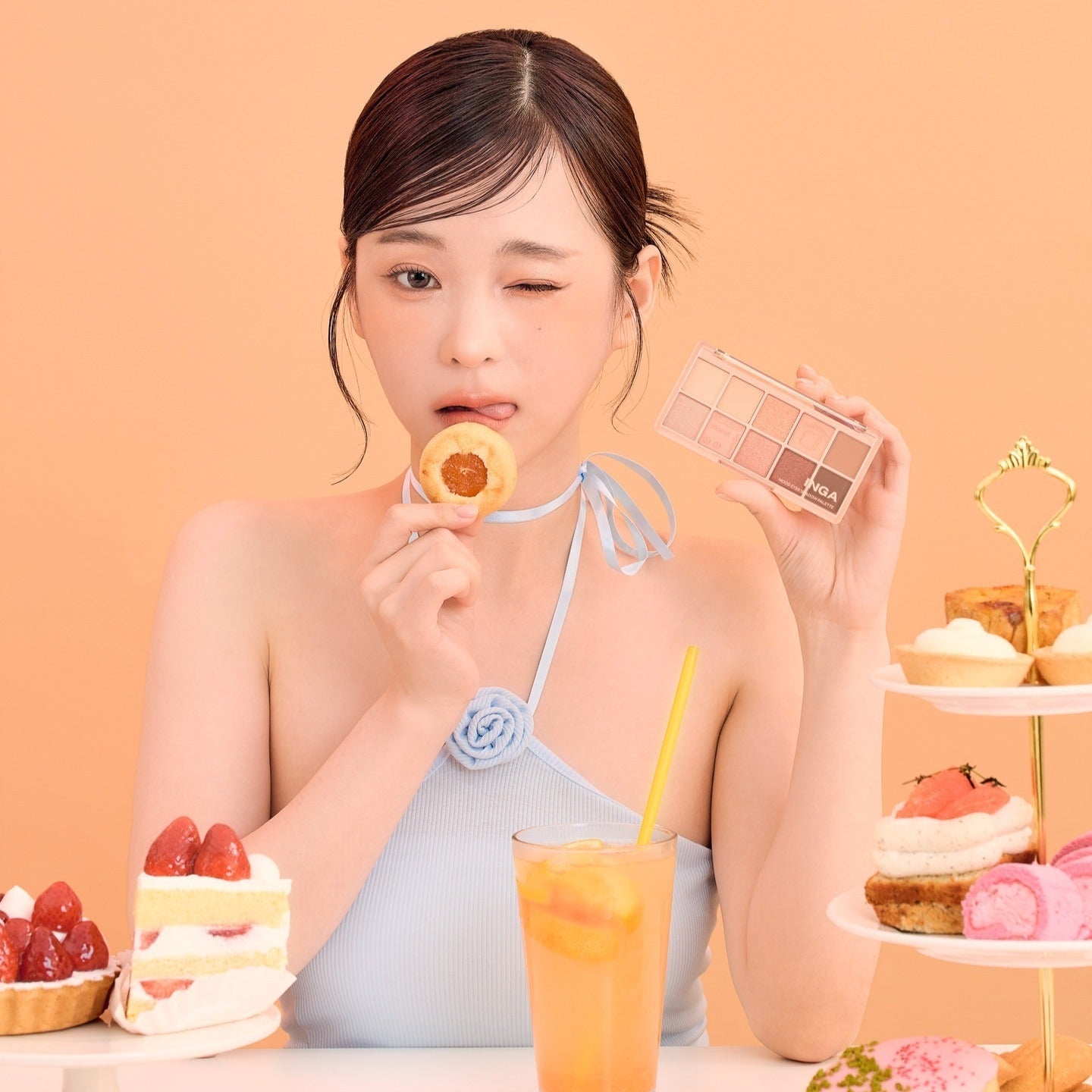 Woman holding INGA Mood Eyes Shadow Palette next to desserts, showcasing elegant makeup and lifestyle scene on an orange background.