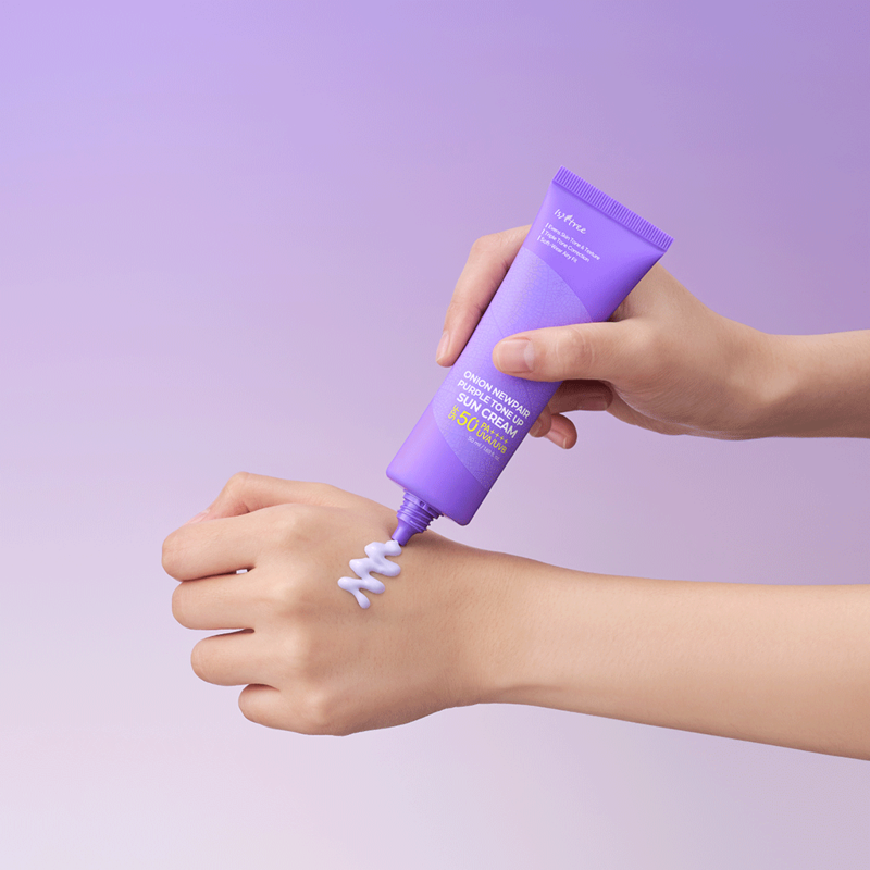 Hand applying ISNTREE Onion Newpair Purple Tone-Up Sunscreen 50ml cream on wrist against a purple background