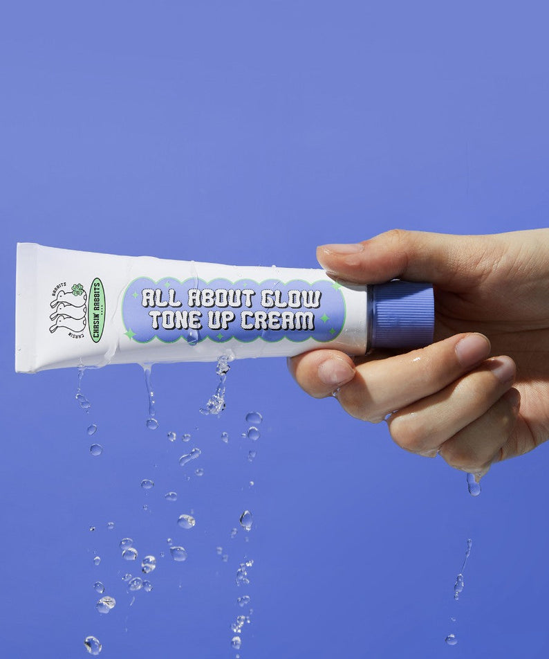 Hand holding IT'S SKIN All About Glow Tone Up Cream against a blue background, with water droplets cascading around the product.