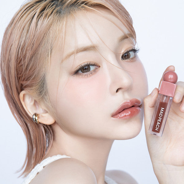 Woman holding IVI MAD PEACH Glow Fit Mood Gloss with elegant makeup look.
