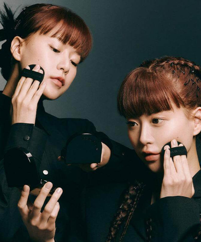 Two models applying JAVIN DE SEOUL Wink Cushion Glow 16g Â, featuring sleek black compacts against a dark background.