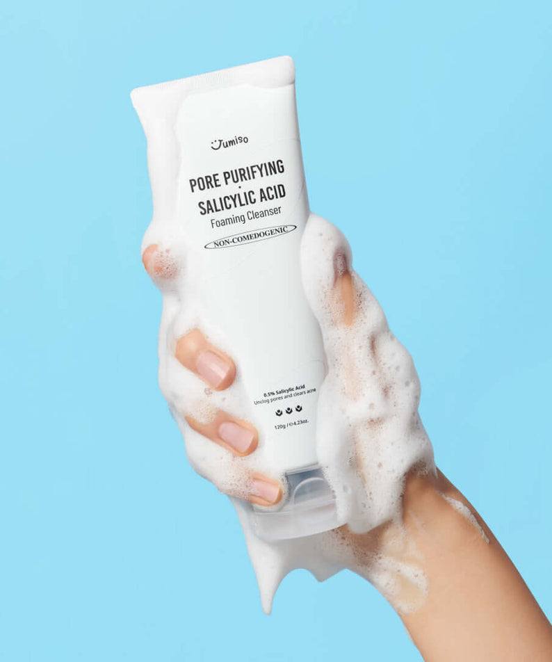 Person holding JUMISO Pore Purifiying Salicylic Acid Foaming Cleanser with foamy bubbles on hand, blue background.