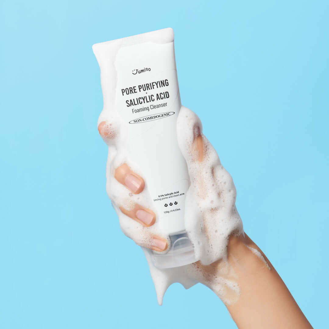 Person holding JUMISO Pore Purifiying Salicylic Acid Foaming Cleanser with foamy bubbles on hand, blue background.