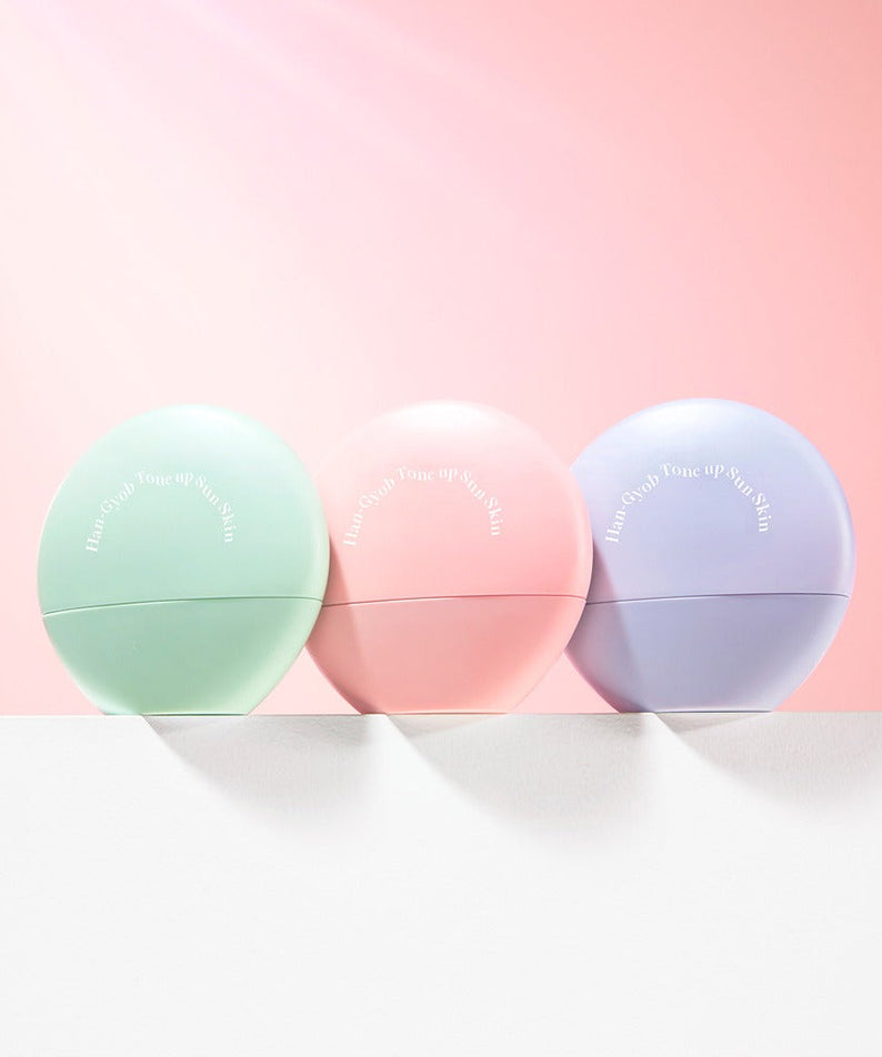 Collection of KAHI One Layer Tone-up Sun Skin 40ml in green, pink, and purple containers against a pink background.