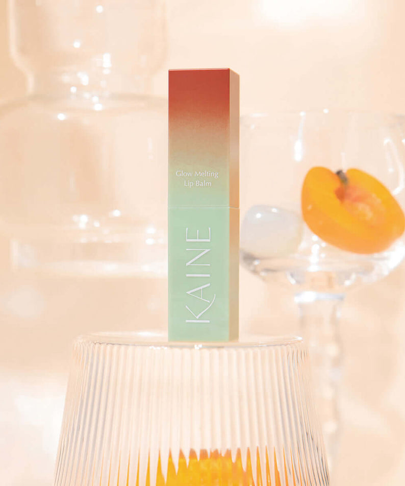 KAINE Glow Melting Lip Balm product on display with decorative glassware background.