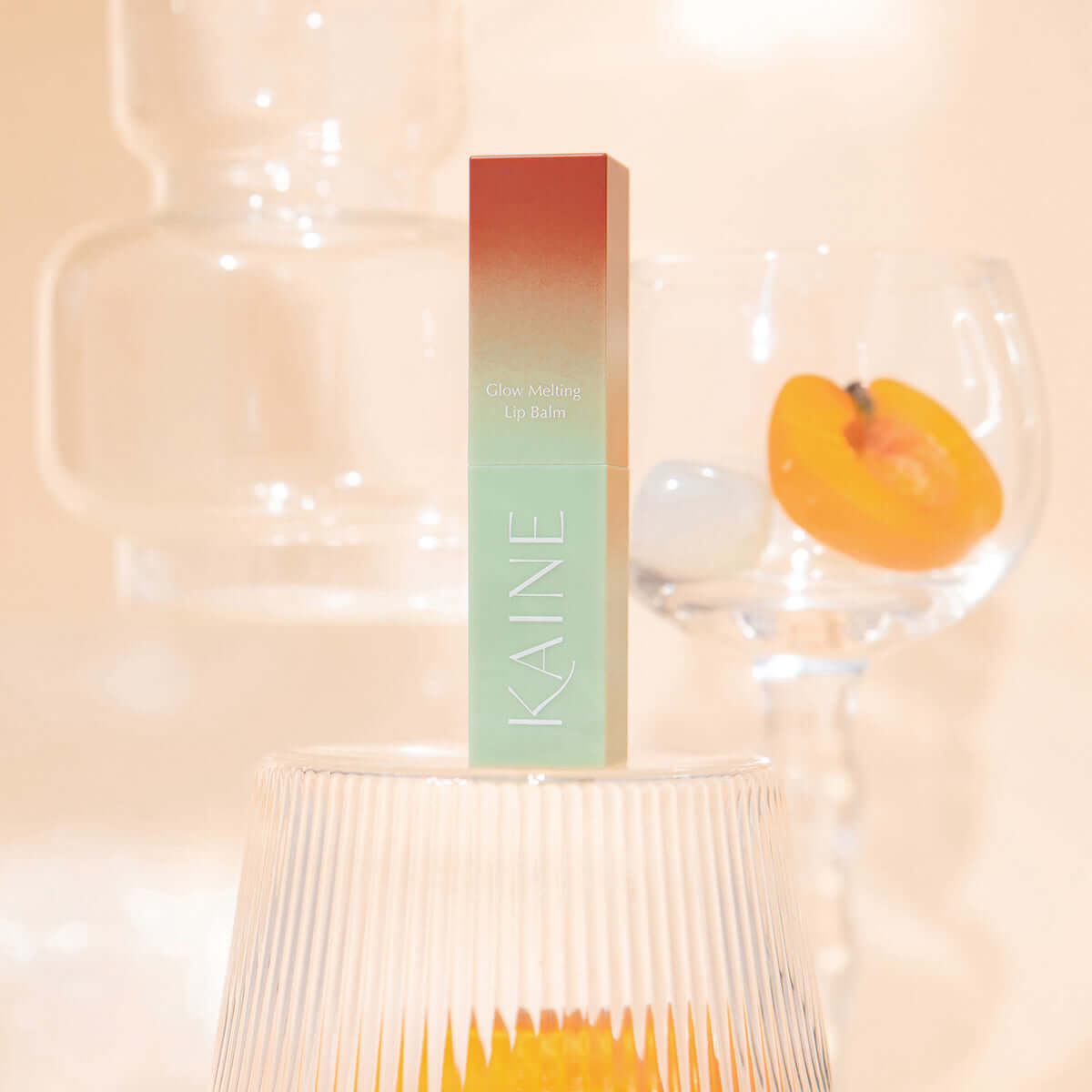 KAINE Glow Melting Lip Balm product on display with decorative glassware background.