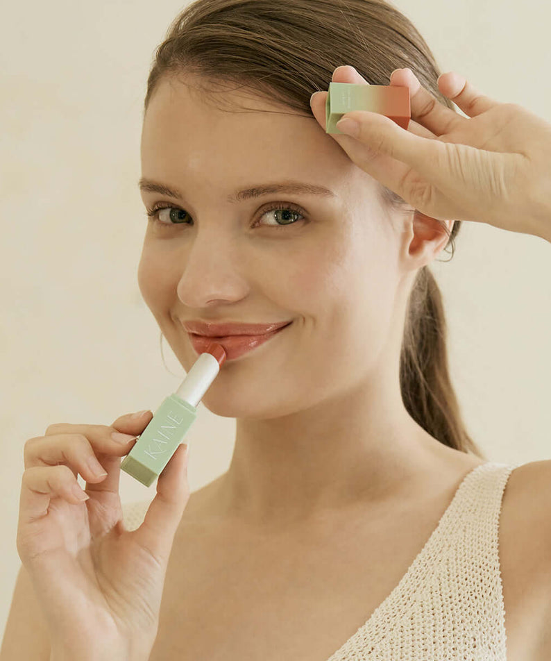 Woman applying KAINE Glow Melting Lip Balm, showcasing the product's packaging and creamy texture for a radiant finish.