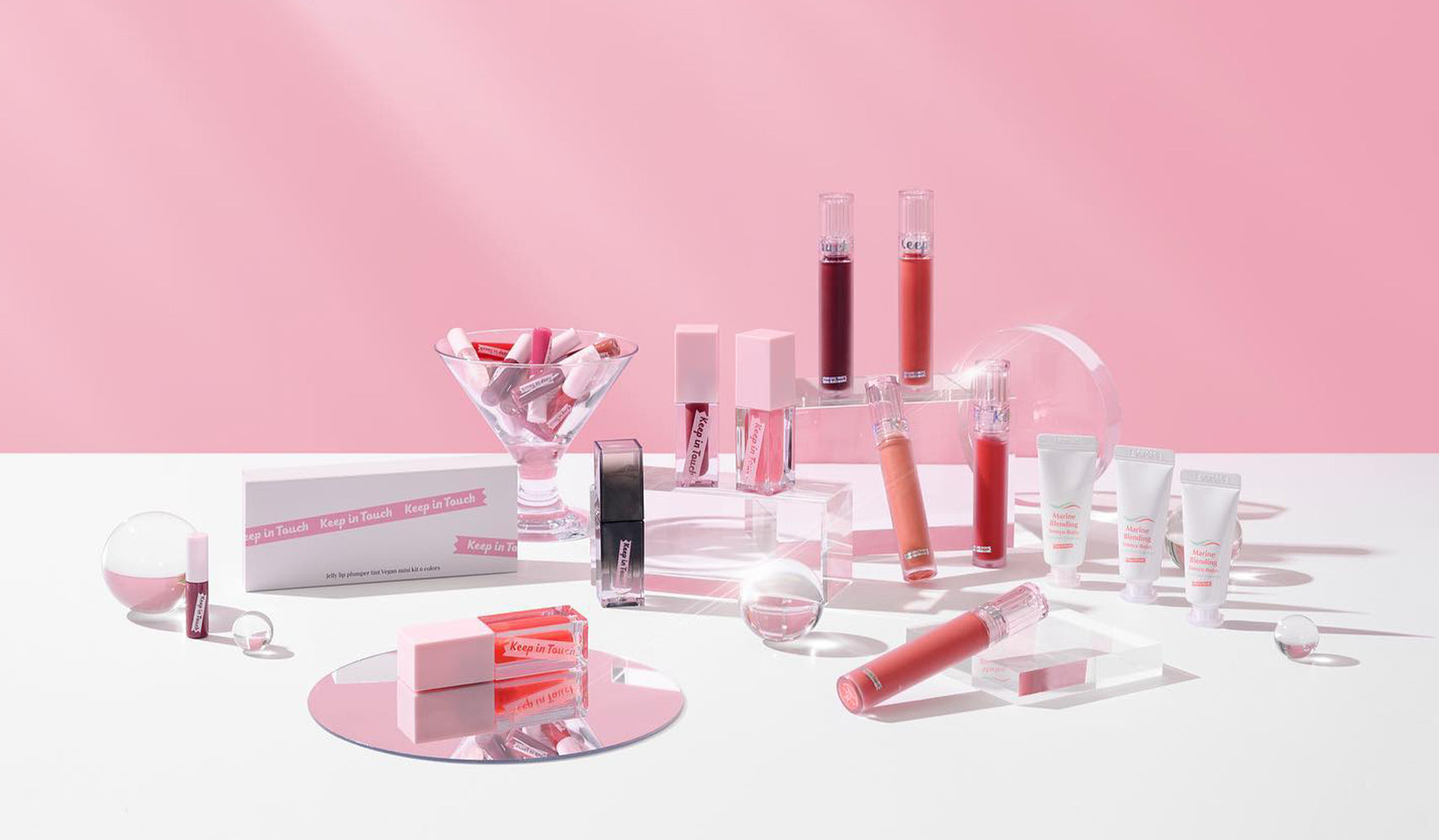 Assorted cosmetic products displayed on pink background, featuring lip glosses, skincare creams, and beauty accessories.