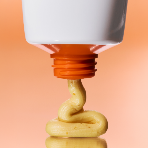KLAIRS Freshly Juiced Vitamin Pack Cleanser 150ml being dispensed showing its creamy texture against an orange background.