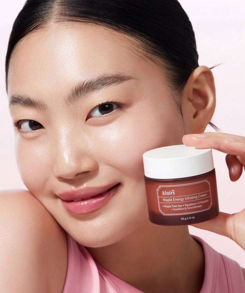 Smiling woman holding KLAIRS Maple Energy Infusing Cream 60g against light background.